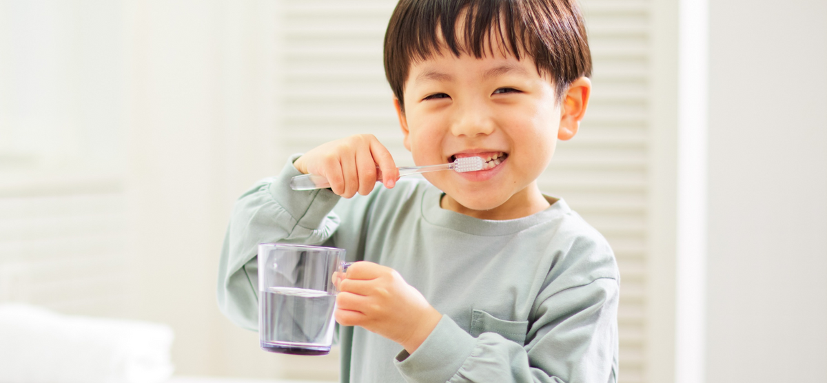 Visuel d'une petit garçon en train de se brosser les dents en souriant avec un verre d'eau à la main